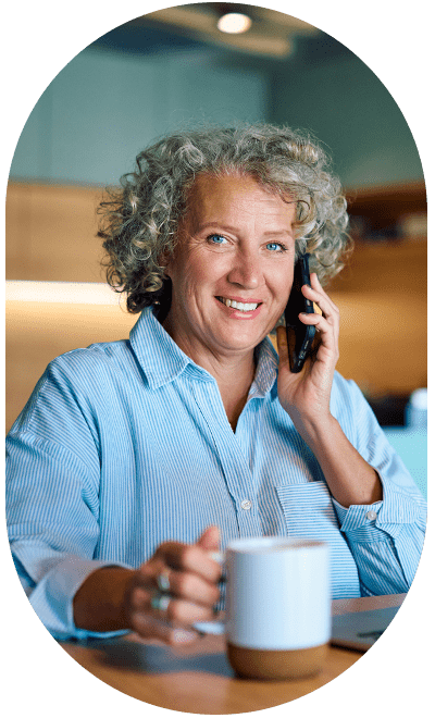 Woman with curly white hair holds a coffee cup while smiling to the camera while using a cell phone